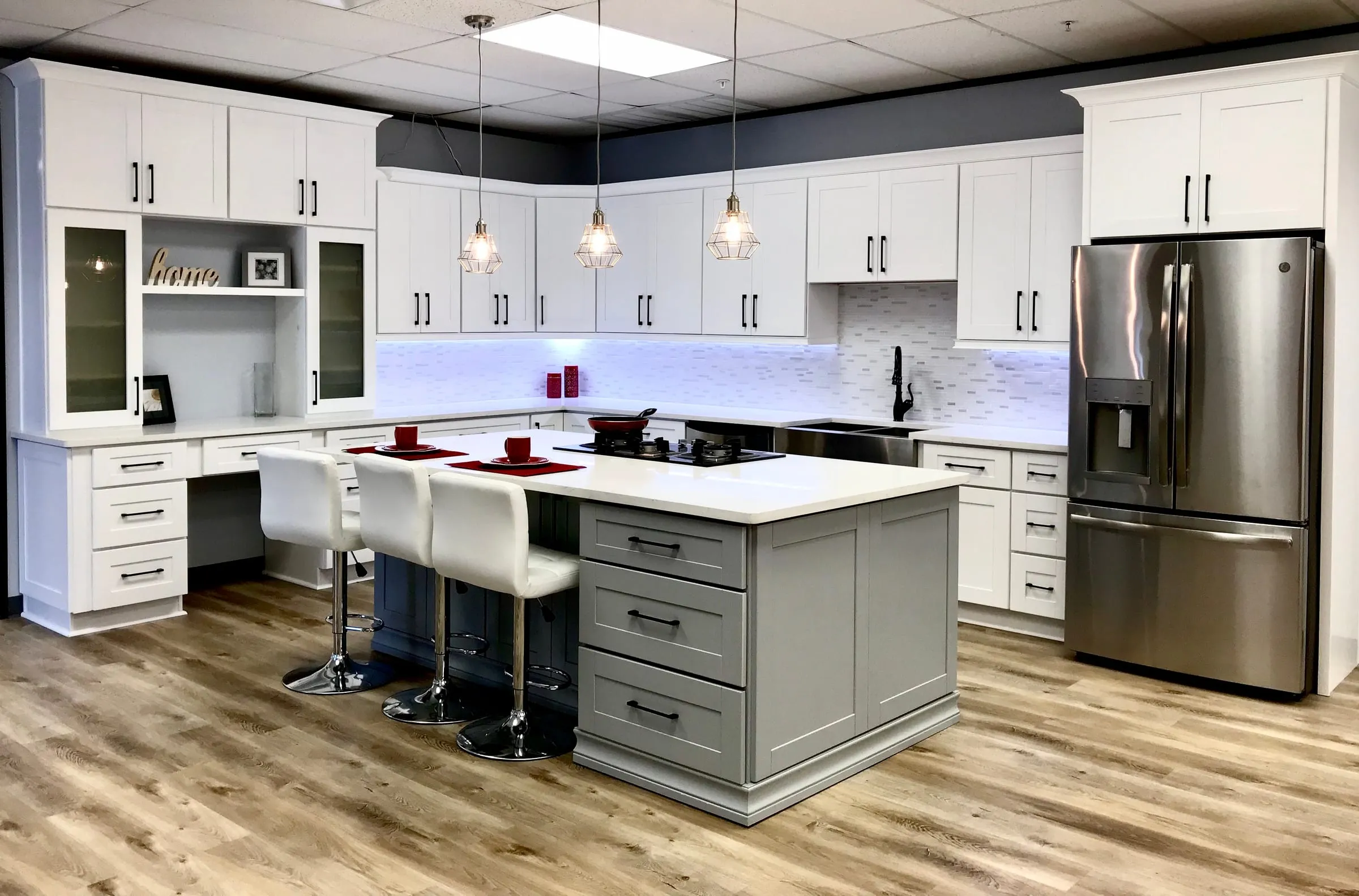 White shaker kitchen with a contrasting gray island, bar stools, glass-front uppers, and under-cabinet lighting
